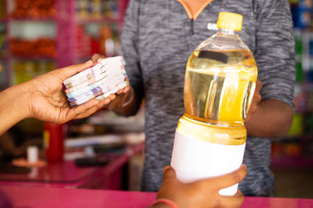 Focus on hand, Close up shot of customer buying edible oil by paying stack or bundle of money at kirana or grocery retail shop - concept of inflation or increase in prices of daily needs and cost of livingの写真素材