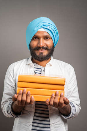 Portrait shot of Young smiling sikh man looking at camera by holding books - concept of education,...