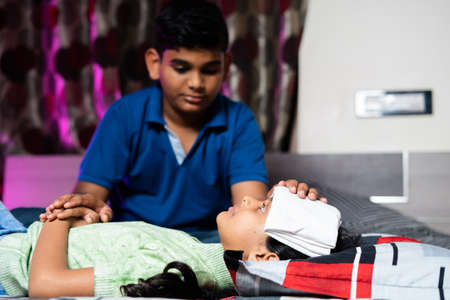 Brother helping by placing cold water cloth on sick sisters forehead due to fever - concept showing...