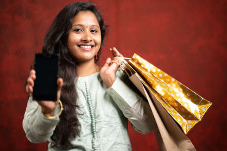 Happy smiling Indian girl with Shopping bags showing mobile phone by looking at camera - concpet of online e-commerce shopping and festival sales.の写真素材
