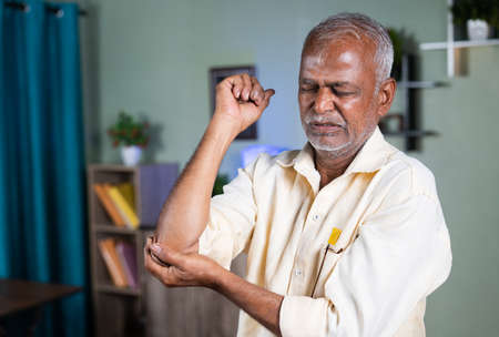 indian senior old man exercising by strenching hand due to elbow joint pain at home - concept of suffering joint pain and sprain injury.の写真素材