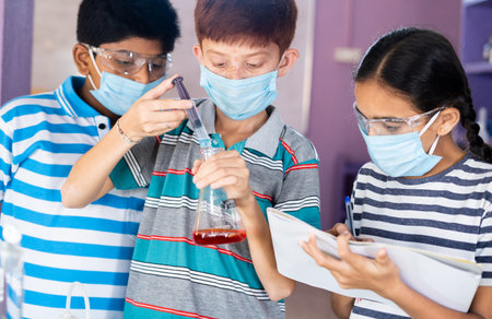 Diverse Kids experimenting with safety eyeglasses and face mask by noting results while mixing chemical using flask at chemistery laboratory - concept of Education, learning and childhood development.の写真素材
