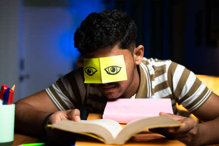 Young student acting like reading while sleeping by putting eye open stickers at home - concept of lazyiness and late night studying, careless and negligence.の写真素材