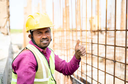 Construction worker with hardhat and jacket showing thumbs-up while working by looking at camera -...