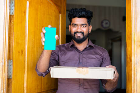 Happy smiling customer with courier box in hand showing green screen mobile phone by looking at camera at home - concept of happy customer service and app promotionの写真素材