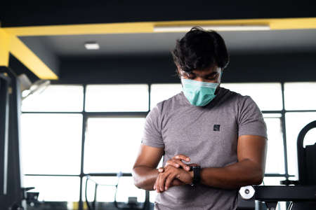 Young muscular builder with medical face mask checking workout plan or monitoring hart rate from smartwatch at gym - concept of healthcare, technology and safety measures during  pandemic.の写真素材
