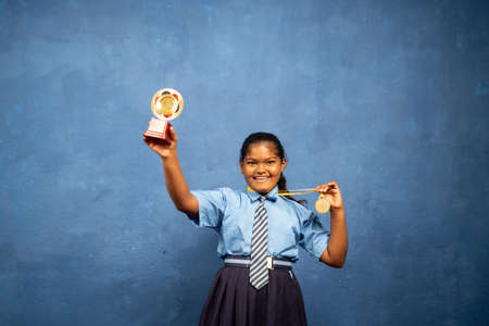 Enjoying indian school girl kid celebrating by showing trophy and medal by looking at camera - concept of achievement, proud, successful.の写真素材