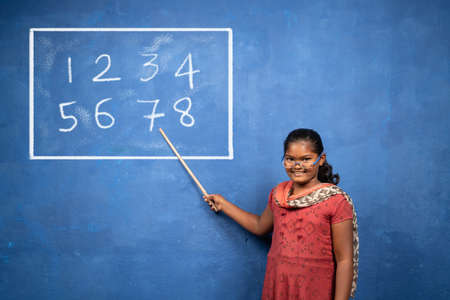 Girl kid with stick in hand pointing to board by acting like teacher or professor - concept of childhood dreams, education and playful lifestyleの写真素材