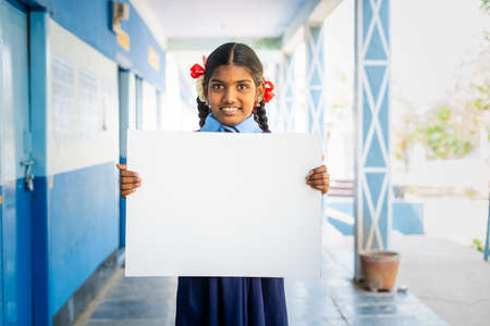 Happy girl kid in uniform showing white empty board or placard by looking at camera at school corridor - concept of education, announcement and learning promotion.の写真素材