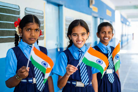Smiling girl kis in unifrom waving Indian falg by looking at camera at school corridor - concept of Patriotism, republic or independence day celebrationの写真素材