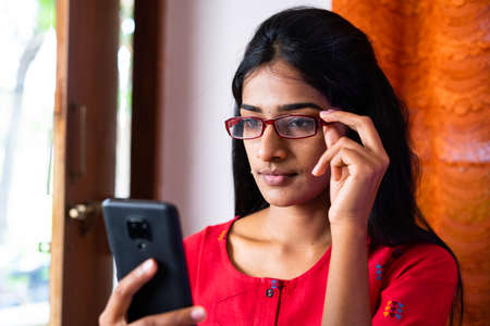 Young girl with eye glasses busy using mobile phone at home - concept of social media, internet and eyesight problemの写真素材