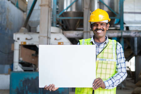 smiling industrial worker with safet hat hosling empty placard or sign board while looking at camera at factory - concept of promotion, advertisement and blue collar jobs.の写真素材
