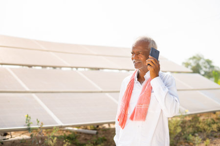 happy Indian farmer talking on mobile phone in front of solar panels about electric power generation farmland - concept of technology and modern agriculture and communicationの写真素材