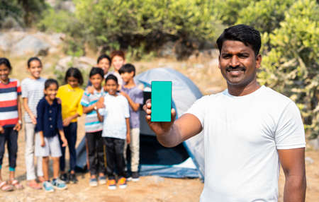 Teacher showing green screen mobile phone by looking camera at summer camp while kids playing background in front of tent - concept of app promotion, advertisement and online booking service.の写真素材