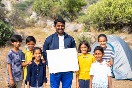 Smiling teacher with teenage kids showing white empty board by looking at camera at mountain top - concept of advertisement, trekking and promotion for summer camp.の写真素材