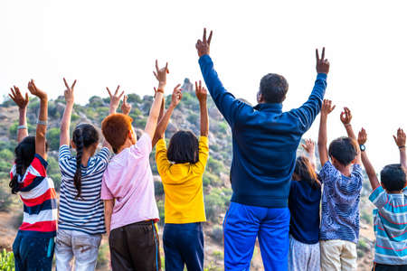 Back view shot of group of teenage kids with teacher celebrating by raising hands on top of hill after reaching destination - concept of summer holiday, trekking, outdoor adventures activities and bondingの写真素材
