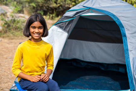 happy Smiling girl kid sitting near camping tent by looking at camera at forest during hot summer vacation - concept of happiness, relaxation and weekend activities.の写真素材