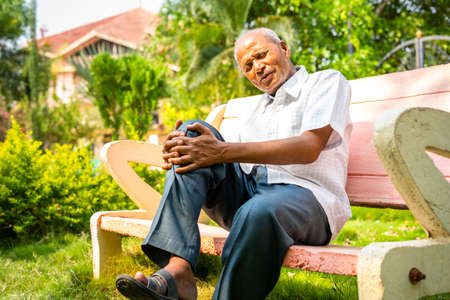 Senior old man suffering from knee joint pain while sitting at park - conept of arthritis, healthcare and injuryの写真素材