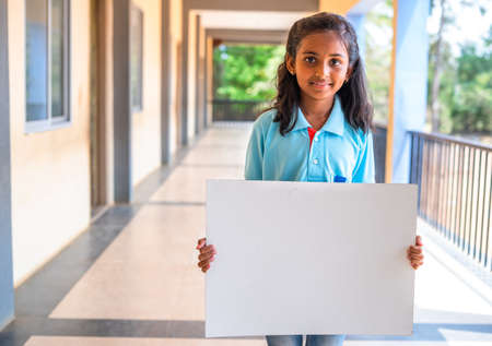 Smiling girl kid showing white empty board by looking camera at school corridor - concept of education, advertisement and promotionの写真素材