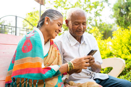 Senior couple using mobile phone while sitting on park - concept of social media, technology lifestyles and relaxationの写真素材