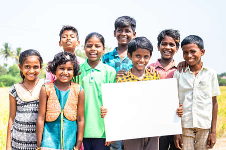 Group of happy smiling Indian village children with one kid holding empty sign board or placard by looking camera - concept of happiness, advertisement and promotion.の写真素材