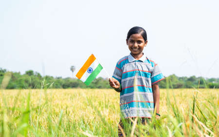 Happy smiling Indian village girl with Indian flag standing in middle of the paddy field by looking...