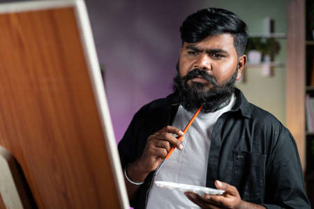 young indian beard artist thinking while painting on canvas board at home - concept of hobbies, leisure activity and professional artist.の写真素材