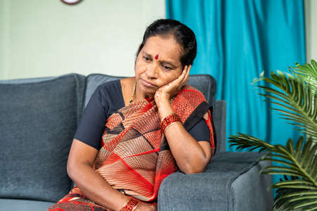 Deep thinking senior woman while sitting on sofa at home - concept of loneliness, upset and worried.の写真素材