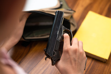 Close up shot of kid taking out gun from back pack at school on desk with books - concept of US or...