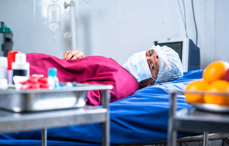 Senior sick patient on hospital bed with medical face mask and operation gown looking at camera -...