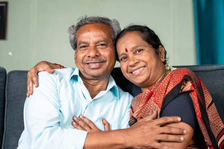 Happy smiling senior Indian couple embracing each other by looking at camera while on sofa - concept of family bonding, relaxation and happinessの写真素材