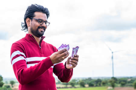 Happy smiling man counting money in front of wind turbine or power plant - concept of saving money...