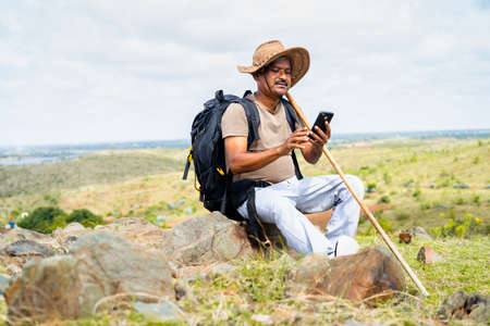 Middle aged with hatusing mobile phone while sitting on top of hill during trekking - concept of internet connection, technology and social mediaの写真素材