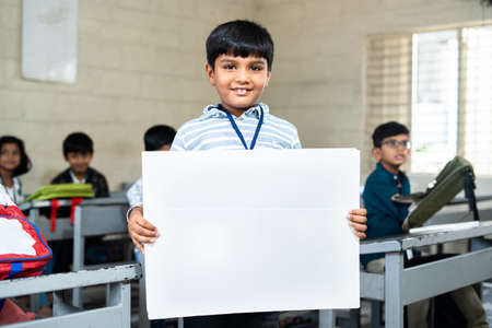 Happy schoolkid showing white empty board by looking camera at classroom in front from to students -...