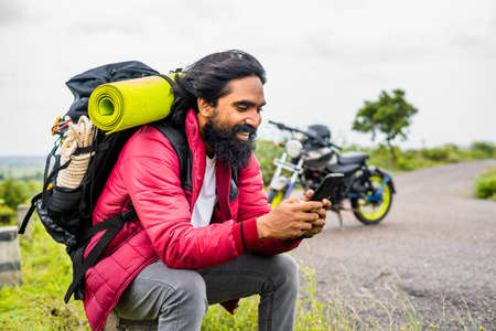 Happy smiling traveler busy using mobile phone while sitting on road side infrtont of motor bike - concept of technology, social media and taking break.の写真素材