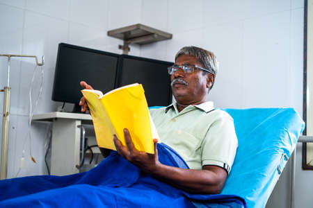 Senior man with eyeglasses reading book at hospital while on bed - concept of relaxation, rehabilitation and medicare treatment.の写真素材