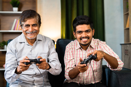 Happy smiling grandfather and grandson playing video game using game pad by looking camera at home -...