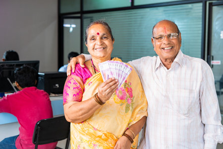 Happy smiling senior couple showing indian money or currency notes by looking camera at bank -...