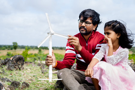 Father playing and explaining to daughter by holding toy wind power fan on top of mountain - concept of development, bonding and renewable energyの写真素材
