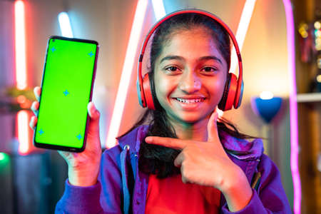 Happy teenager girl kid with headset showing green screen mobile phone by pointing finger by looking camera at home - concept of app advertisement, entertainment and promotion.の写真素材