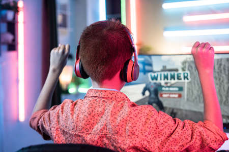 Back view shot of teenager kid celebrating victory by dancing on chair while playing video game on computer at home - concept of achievement, positive emotion and excitement.の写真素材