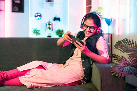 excited girl playing video game on mobile phone with wireless headset while sitting on sofa at home - concept of relaxation, hobby and entertainment.の写真素材