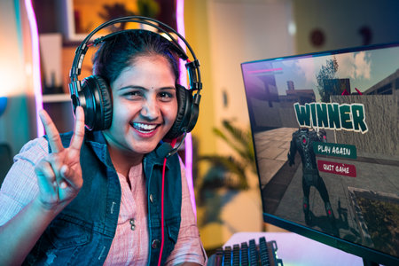 Smiling gamer with headphones celebrating win game by showing victory sign while playing live video game on computer at home - concept of championship, tournament and achievement.の写真素材