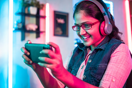 happy smiling girl with headphones and eyeglass playing online video game on mobile phone at home - concept of live streamer, holydays and cyberspaceの写真素材