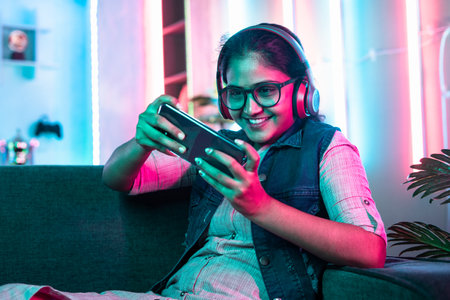 excited girl playing video game on mobile phone with wireless headset on sofa at home on neon background - concept of cyberspace, hobby and entertainment.の写真素材