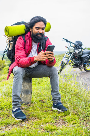 vertical shot of Happy smiling traveler busy using mobile phone while sitting on road side infrtont...