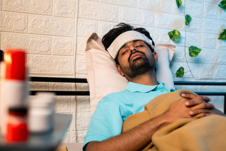young indian sick man suffering from mild fever while sleeping at night by placing cold cloth on forehead - concept of healthcare, covid symptoms and illness.の写真素材