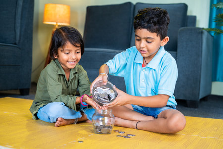 Young sibling kids putting coin on glass while sitting on floor at home - concept of saving money, togetherness and future educationの写真素材