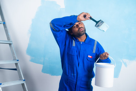 Tired painter worker while painting on wall by holding paint bucket at workplace - concept of overworked, hard working and maintenance service.の写真素材