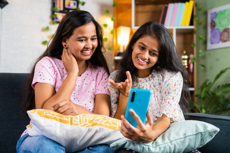 Happy smiling sibling girl sisters on sofa making video call on mobile phone at home - concept of relationship, wireless communication and cyberspace.の写真素材